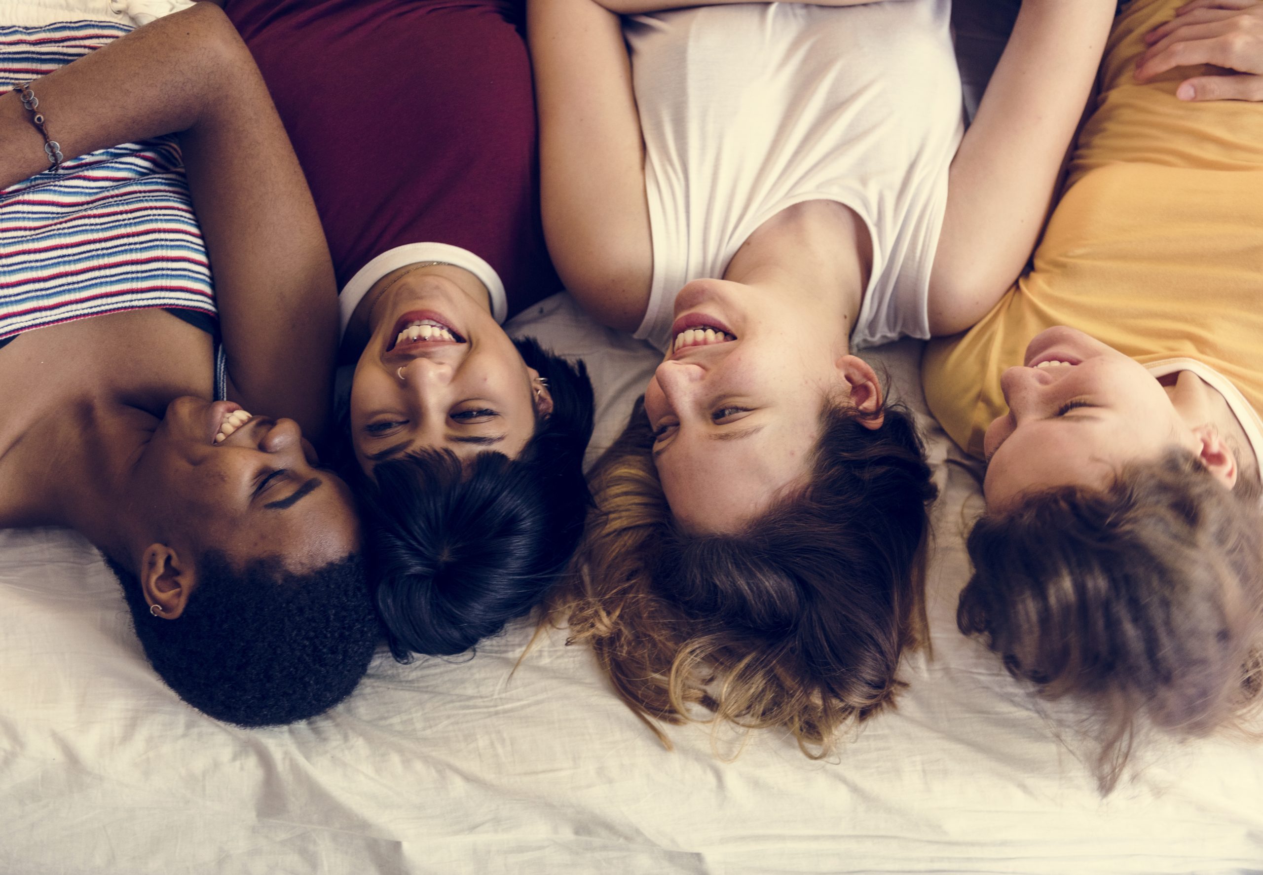 A group of four diverse women lying on a bed together. They are all laughing.