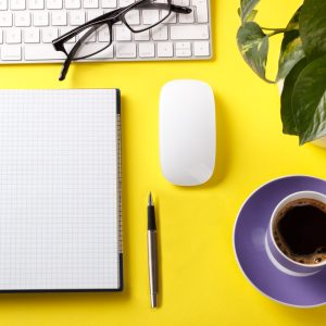 A top view of a modern yellow desktop with a keyboard, notepad, pen, plant, glasses, and a cup of coffee on top.