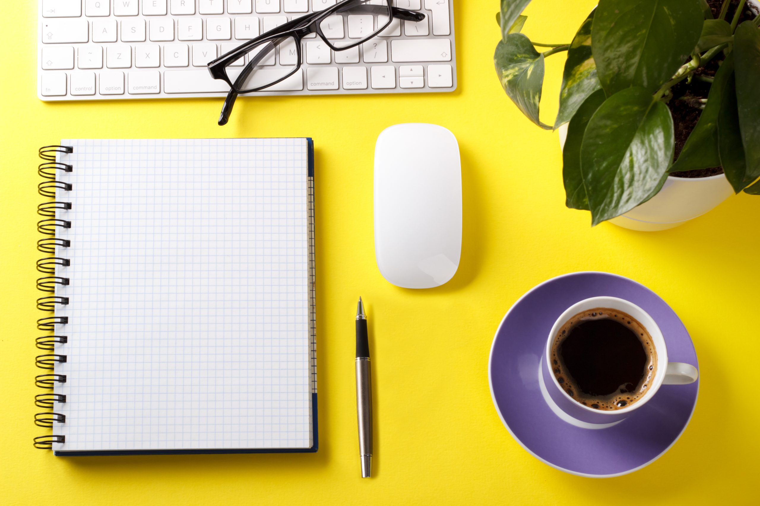 A top view of a modern yellow desktop with a keyboard, notepad, pen, plant, glasses, and a cup of coffee on top.
