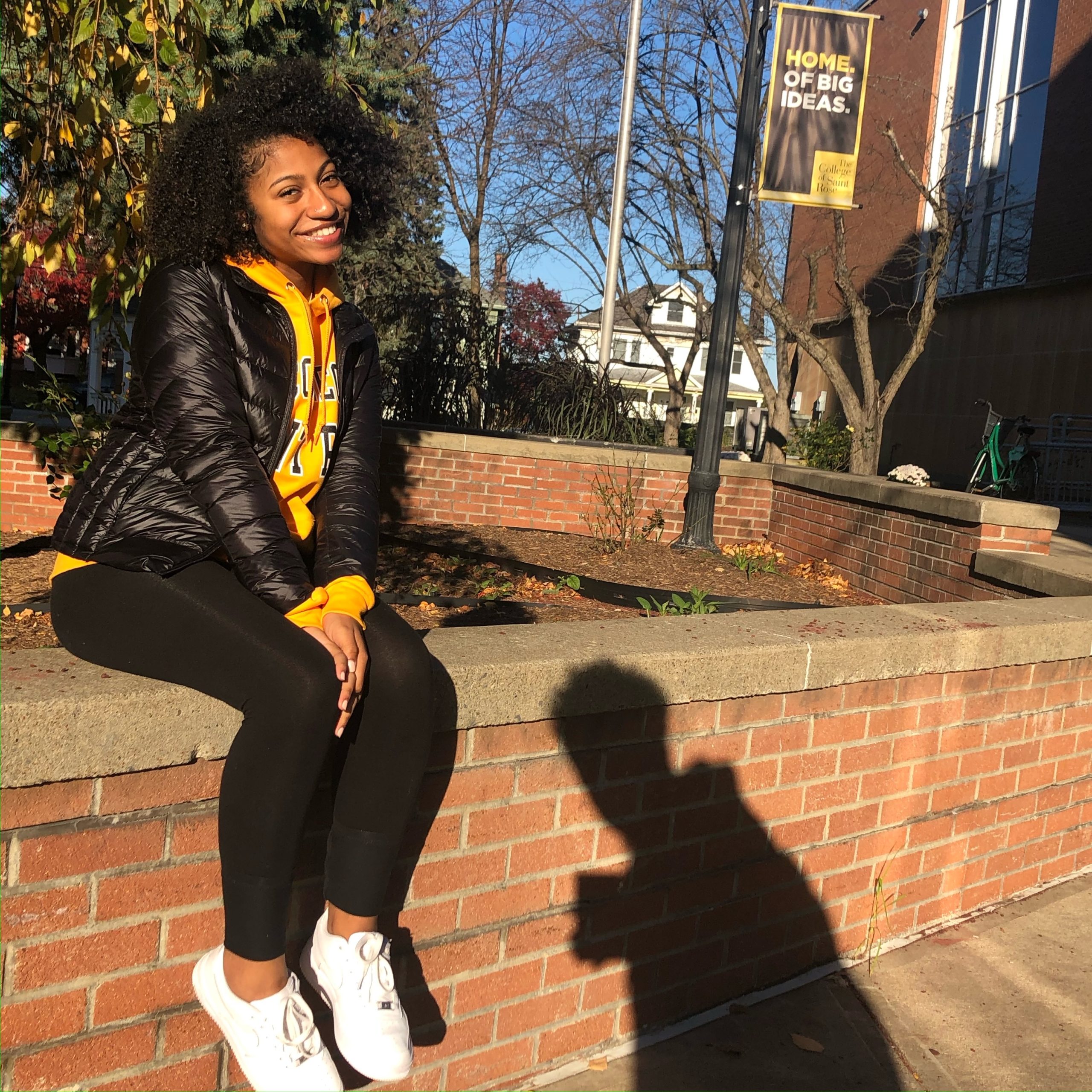 A female student sits on a brick half wall on the Saint Rose campus on a sunny day. She is smiling and wearing a Saint Rose sweatshirt under a black jacket.