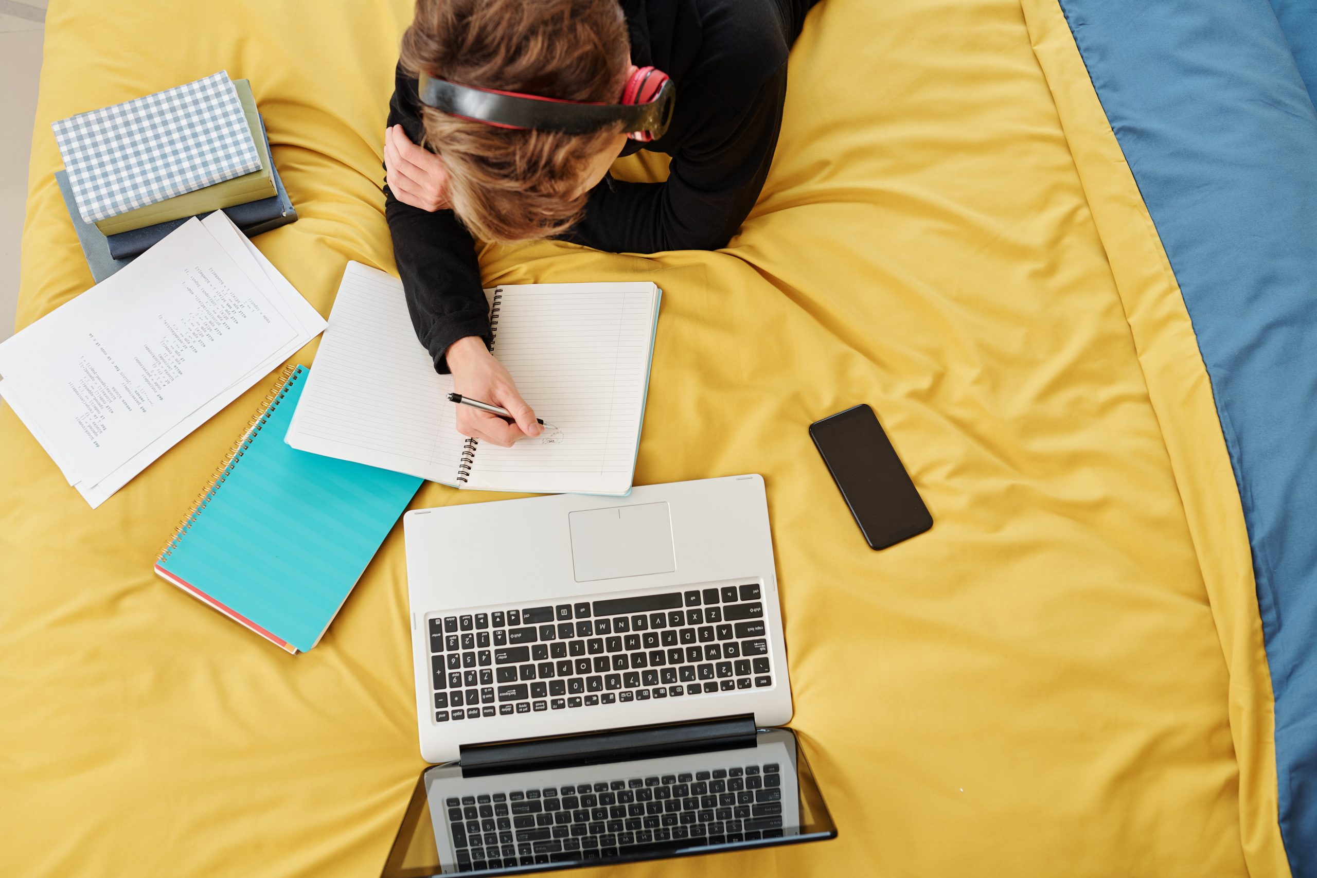A student laying on a yellow blanket while drawing and writing in a notebook. There are an open computer, phone, and other notebooks around them.